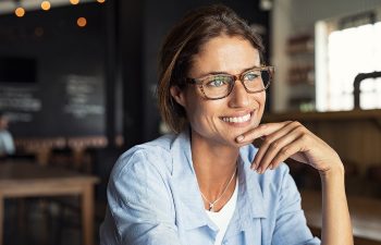 A woman wearing glasses and a light blue shirt smiles while sitting indoors with blurred background elements.