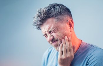 A man with short hair holds his face in pain, eyes closed, wearing a blue shirt against a light blue background.