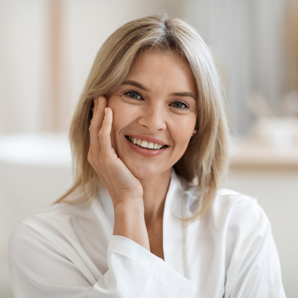 A woman with blonde hair smiles, resting her face on her hand. She is wearing a white shirt and the background is softly blurred.